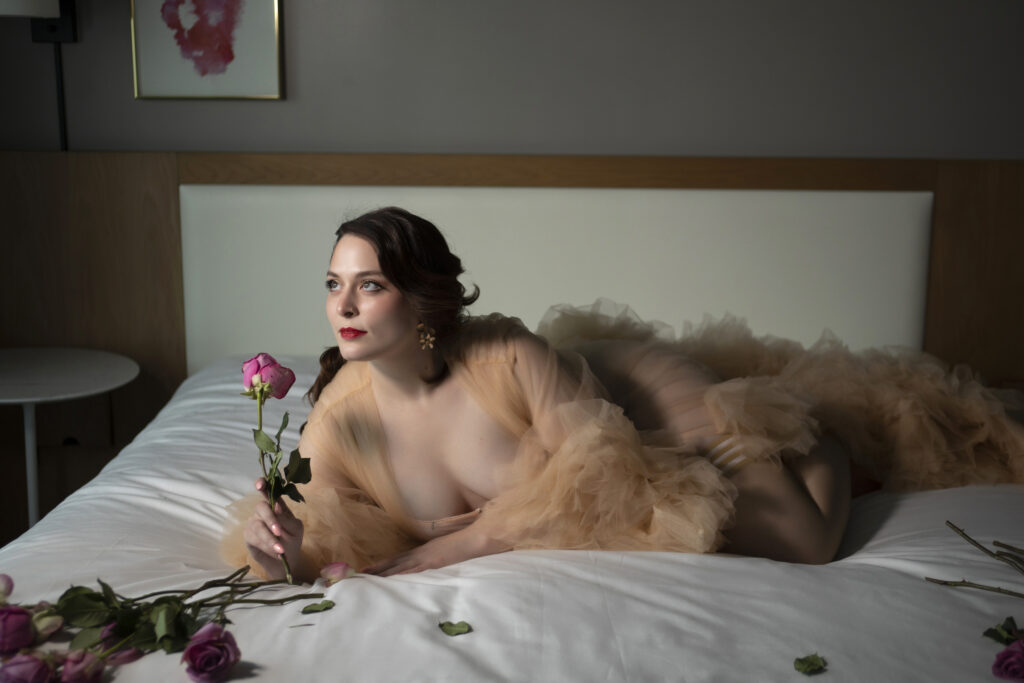 A woman posing on the bed with a gold robe and holding a pink rose to her nose.